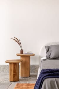 A modern minimalist bedroom with a white bed and wooden side tables and stools. The room features a vase with dried flowers o