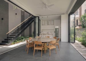 A modern residential dining room featuring a round marble table surrounded by wooden chairs, with a staircase and greenery in