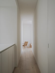 A narrow hallway with white walls and wooden flooring, featuring a single wooden chair in the distance.