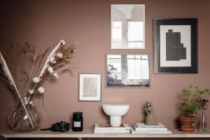 A minimalist home library with a ceramic bowl, books, and plants on a wooden surface against a mauve-colored wall with framed