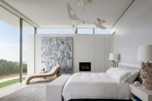 A minimalist bedroom with modern white furniture, large windows, and abstract artwork.