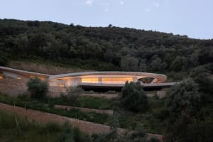 The image depicts a modern residential house with a circular design and concrete material, surrounded by a natural landscape 