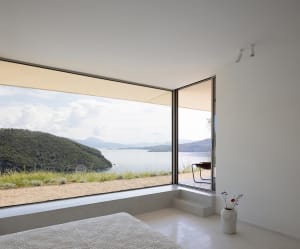 Minimalist bedroom featuring concrete flooring and walls, with large windows offering scenic views of a coastal landscape.