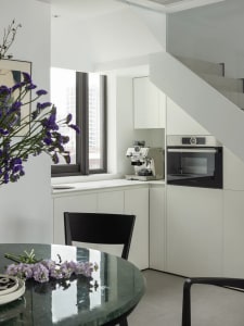 A modern kitchen featuring white cabinets, a black marble table with purple flowers, and a window with a cityscape view.