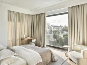 Modern minimalist bedroom featuring a large window with cityscape view, neutral textiles, and sleek furnishings.