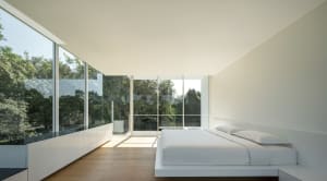 Minimalist bedroom with white furniture, large glass windows, and a view of green trees.