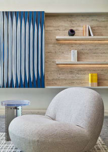 A minimalist living room with a gray upholstered chair, a transparent side table, and a blue and white abstract painting on a