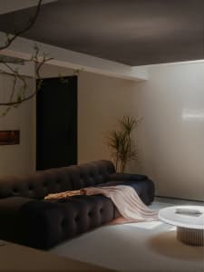 Modern living room with textured cloth sofa, round glass table, potted plant, and white walls.