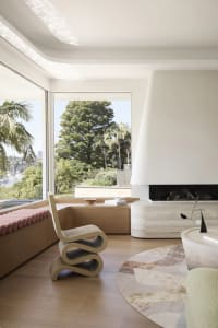 Modern living room with a wooden floor, white fireplace, and large windows overlooking greenery.