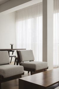 A modern minimalist living room featuring upholstered fabric chairs and wooden tables with sheer curtains allowing natural li
