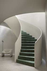Spiraling staircase with green carpeted steps and white concrete walls.