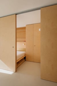 A minimalist bedroom with light wooden walls and paneling, featuring a bed with a storage base and a closet with sliding door