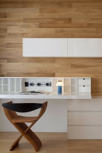 A modern home office featuring a wooden chair and a white desk with a blue lamp on it, set against a wooden wall with built-i