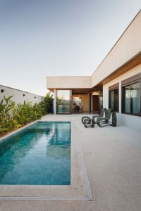 Modern residential backyard featuring a concrete patio, glass patio doors, wooden chairs, and a swimming pool surrounded by l