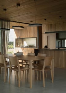 A modern dining room with a wooden table and chairs, featuring large windows and a kitchen counter.