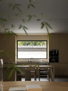 Modern kitchen with wooden cabinets, table, and chairs under a large window overlooking greenery.