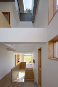 Modern interior with white walls, wooden stairs, concrete ceiling, and large windows showing a green field.