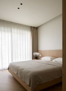 Modern minimalist bedroom with wooden furniture, white bedding, and sheer curtains.