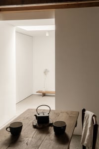 A minimalist interior featuring a wooden table with a teapot and two cups, set in a well-lit room with white walls.