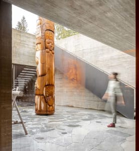 A modern indoor space featuring concrete decoration with a wooden totem pole, stone floor tiles, and a person walking on the 