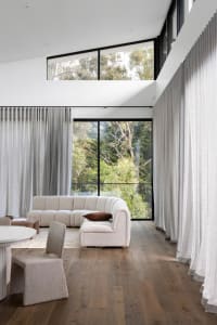 Modern minimalist living room with white fabric sectional sofa, wood flooring, and large windows overlooking greenery.