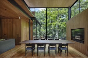 Modern dining room with wooden table and chairs, large windows overlooking greenery.