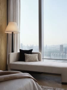 A modern, minimalist living room with a beige sofa and black and textured pillows, featuring a glass wall overlooking a citys