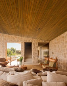 A modern living room with stone walls, wooden ceiling, leather chairs, and glass coffee table.