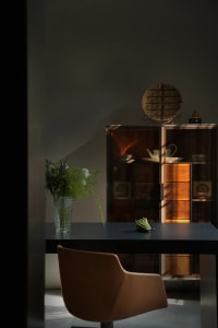 A minimalist desk setup featuring a glass vase with greenery, a leather chair, and a glass cabinet filled with items.