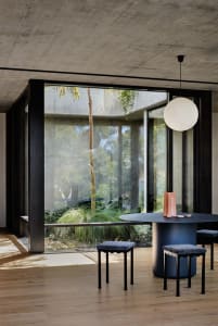 A modern, minimalist dining room with a large round table, stools, and a concrete and glass structure visible through large w