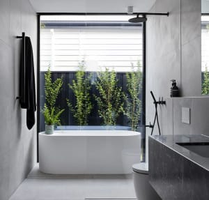 Modern minimalist bathroom with concrete walls, white bathtub, and green plants in front of a black wooden fence.