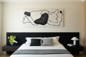 A minimalist bedroom with a textured wooden headboard and white bedding. A black and white abstract painting hangs above the 
