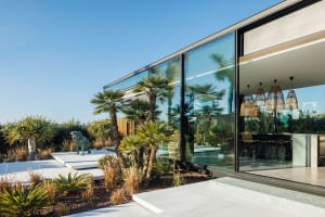 Modern residential house with large glass windows and a landscaped garden on a sunny day.