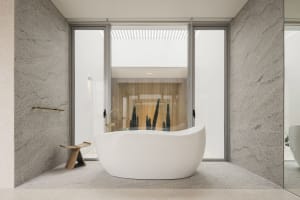 A minimalist bathroom with a white bathtub, wooden stool, and large windows showing a cactus plant.
