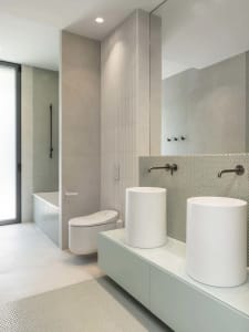 Modern minimalist bathroom featuring a white toilet, white pedestal sinks, and ceramic tiled flooring and shower wall.