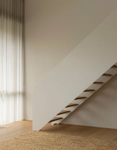 Modern minimalist wooden staircase against a plain wall with a light-colored rug on the floor.
