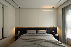 A modern bedroom with minimalist design, featuring a bed with grey bedding and black headboard, illuminated by a linear light