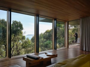A minimalist living room with large windows overlooking a serene coastal landscape, featuring wooden furniture and a yellow s
