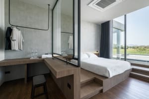Modern minimalist bedroom with white bedding, dark wood flooring, and large windows showing a scenic view.