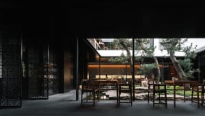 Modern restaurant courtyard with wooden tables, glass walls, and stone elements.