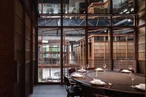 A modern restaurant dining room with a large round table and chairs, made of wood and featuring large windows.