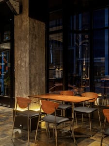 A modern restaurant interior with wooden tables and chairs, glass windows, and a stone wall.