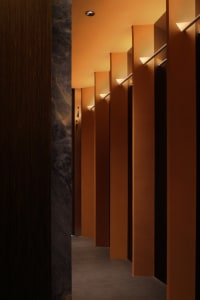 A minimalist hotel corridor featuring concrete walls and floors, with warm lighting and geometric light fixtures.