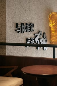 The image shows a wall art piece consisting of metallic Chinese characters displayed on a textured beige wall.