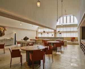 A spacious, modern restaurant interior featuring concrete tables and chairs with high ceilings, pendant lights, and arches.