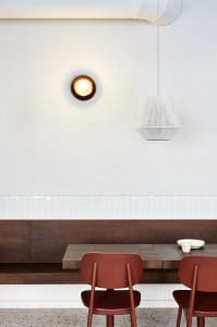 A minimalist cafeteria with wooden furniture and white walls, featuring hanging lights and a built-in bench and table.