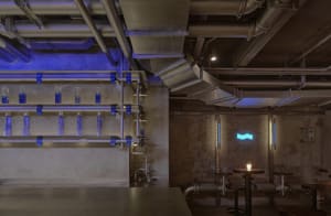 An industrial-style restaurant interior with metal pipes, concrete walls, and blue lighting.