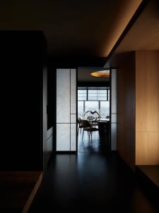 Modern minimalist hallway with wooden walls and a glass partition, leading to a dining area with a round ceiling light and a 