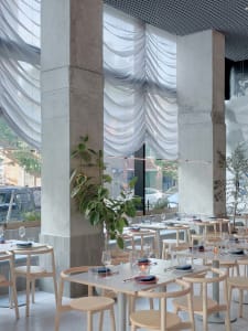 Modern minimalist restaurant interior with concrete walls, glass window treatments, and wooden tables and chairs.