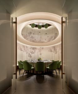 Modern restaurant with a sculptural ceiling and green chairs surrounding a round table set for dining.